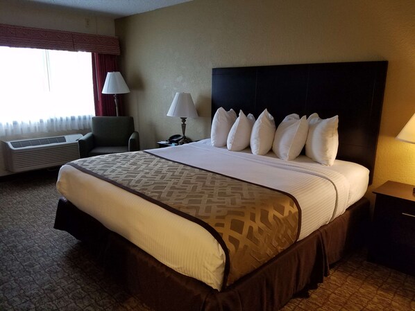 Habitación de hotel con una cama grande, una mesita de noche, una lámpara, una silla y un ventanal con cortinas.