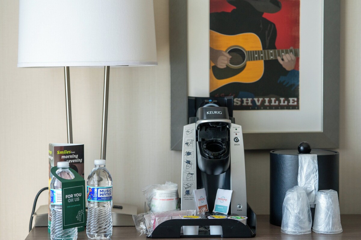 A hotel room with a coffee maker, water bottles, a coffee pod, and a poster on the wall.
