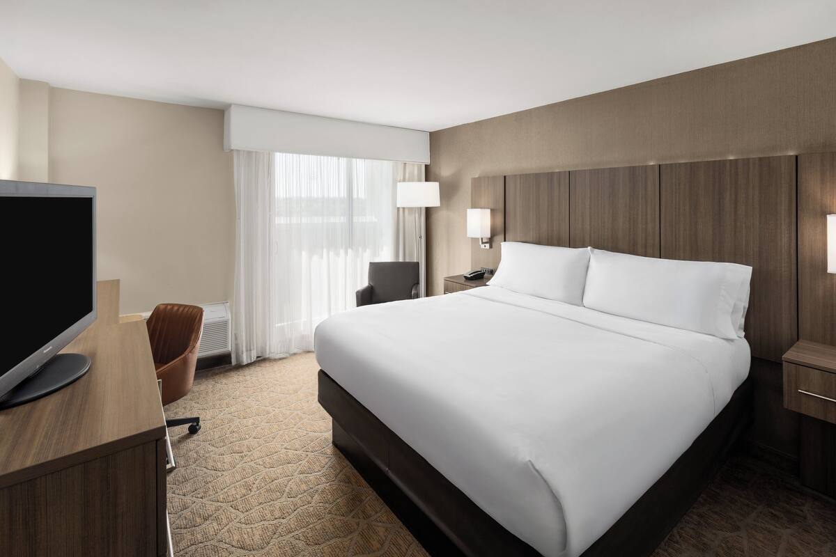 Een hotelkamer met een groot bed, een bureau met een flatscreen televisie, een stoel, een nachtkastje met een lamp en een raam met dunne gordijnen.