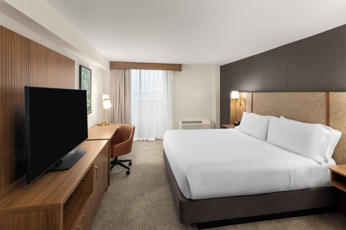 Een hotelkamer met een groot bed, een televisie op een houten bureau, een stoel en een raam met gordijnen.