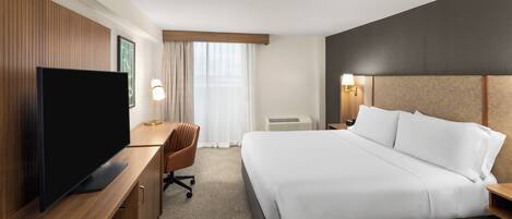 Habitación de hotel con una cama grande, un televisor sobre un escritorio de madera, una silla y una ventana con cortinas.