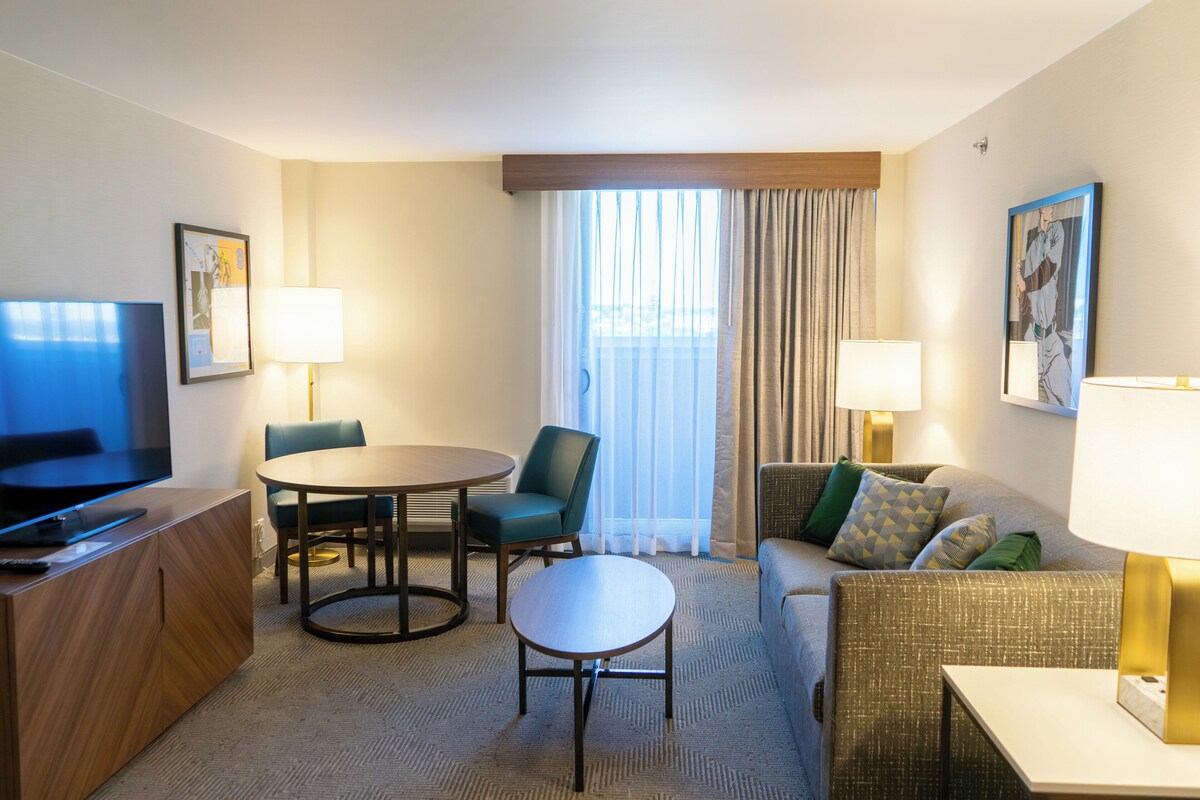 A hotel room with a TV, a dining table with chairs, a sofa, and a window with curtains.