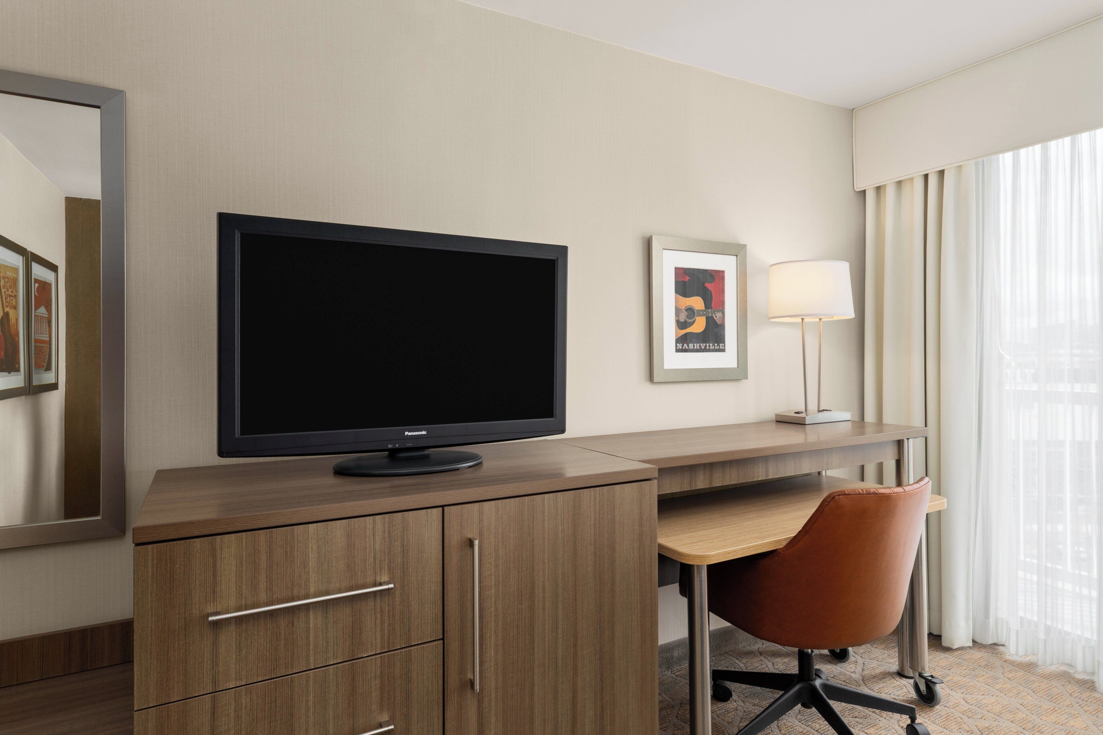 Ein Hotelzimmer mit Flachbildfernseher, Schreibtisch, Stuhl, Lampe und einem fenster mit Vorhängen.