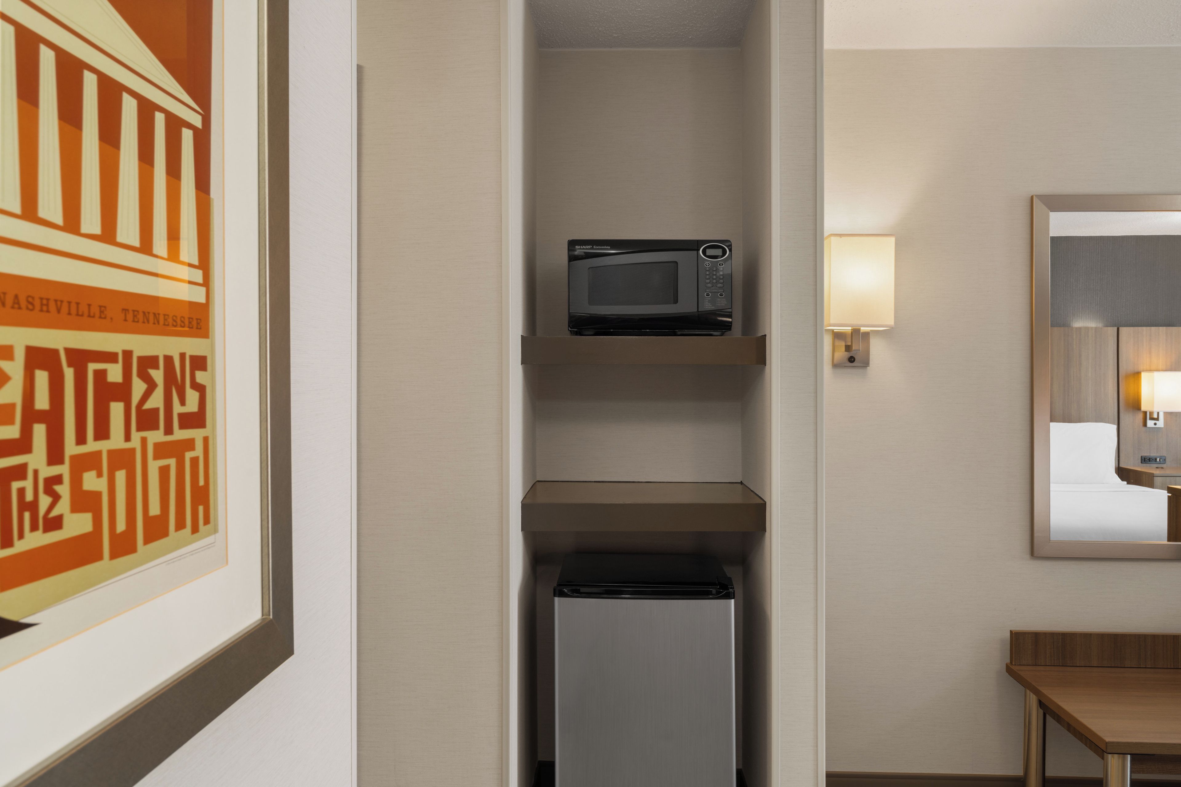 Ein Hotelzimmer mit einer Mikrowelle auf einem Regal, einem Kühlschrank darunter und einem Tisch mit einer Lampe.