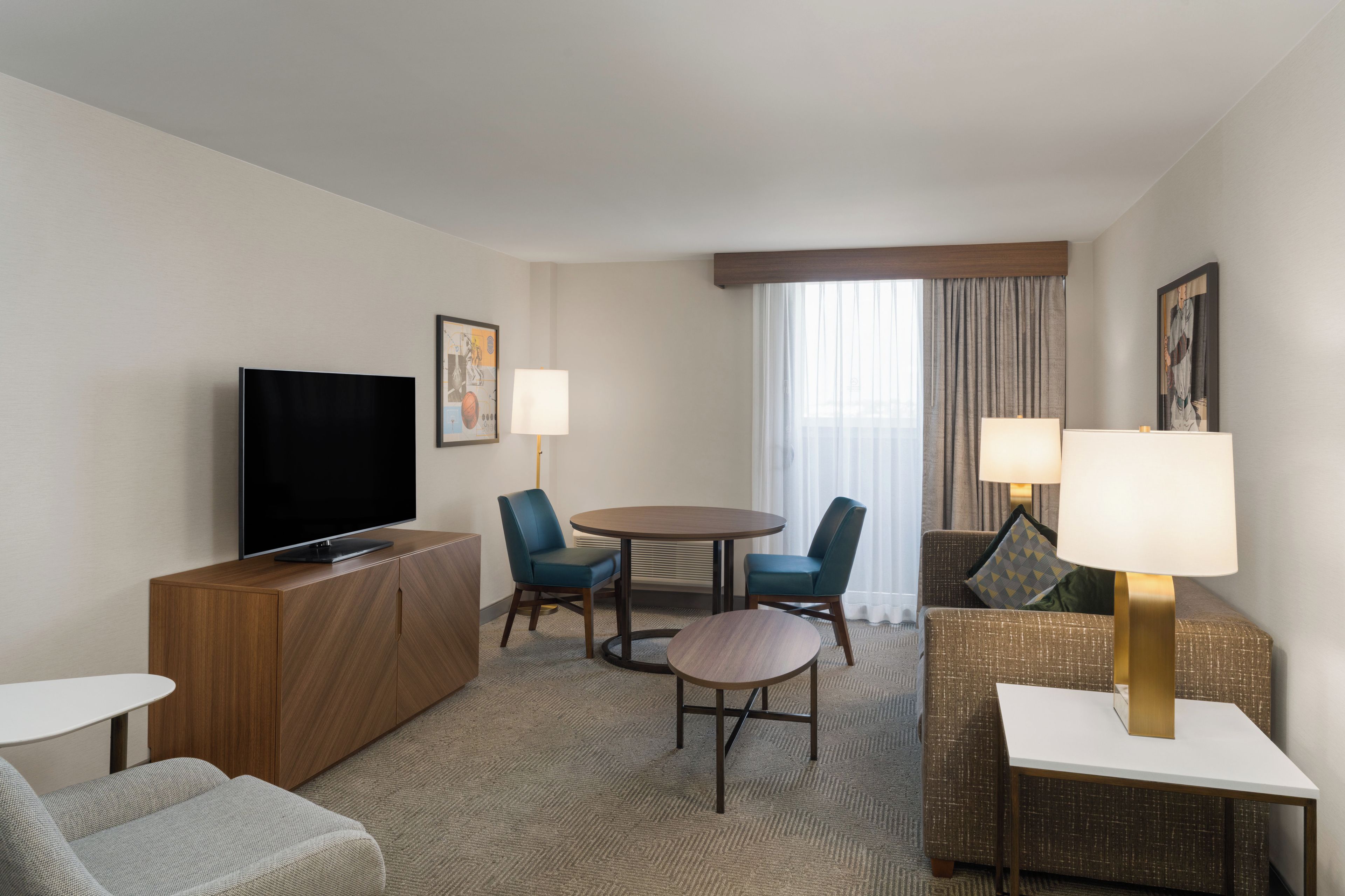 Habitación de hotel con televisor de pantalla plana, una mesa de comedor con sillas, un sofá y una mesita auxiliar con lámpara.