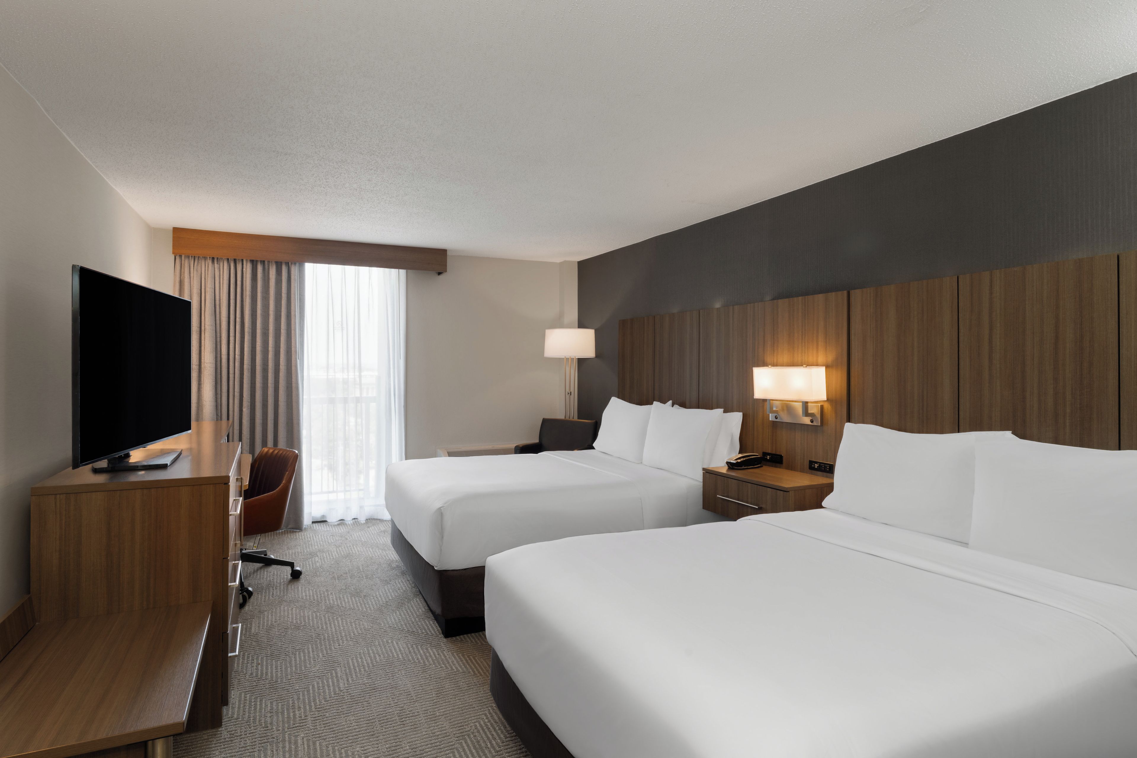 A hotel room with two beds, a desk, a TV, and a window with curtains.
