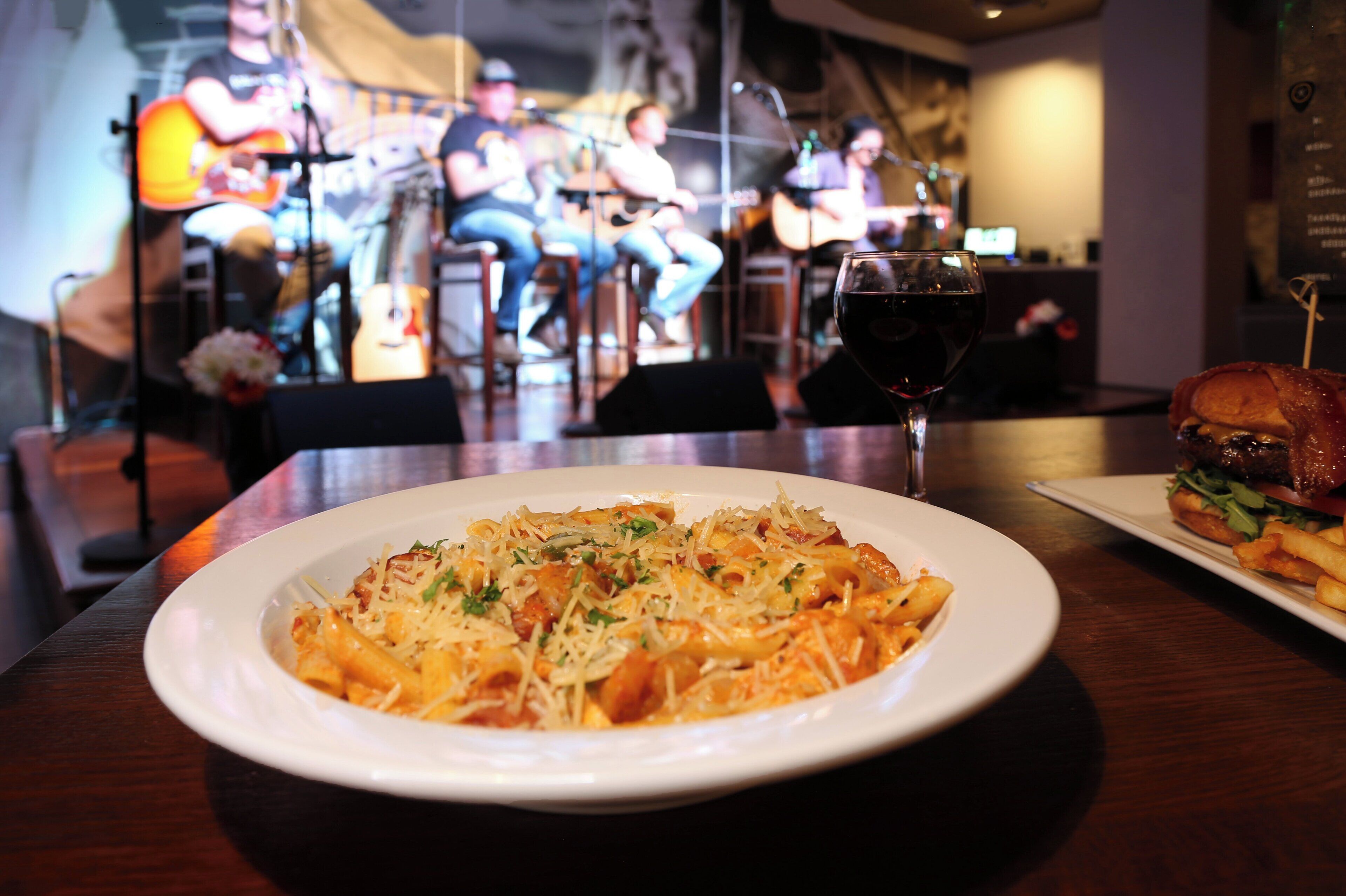 A dining table with a plate of pasta and a sandwich, a glass of red wine, and a band performing in the background.