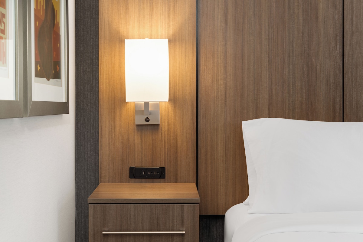 Een hotelkamer met een houten nachtkastje, een wandlamp en een bed met witte beddengoed.