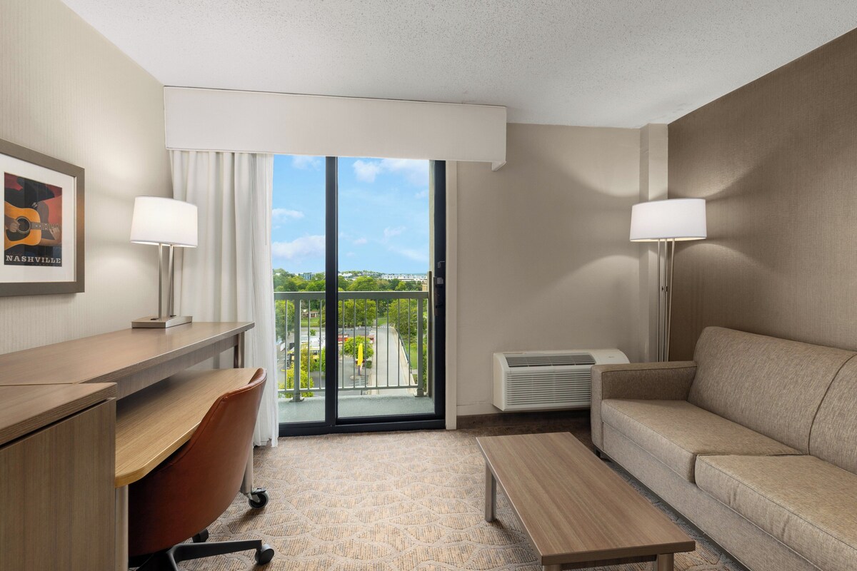 A hotel room with a desk, chair, sofa, and a view of the city through large windows.
