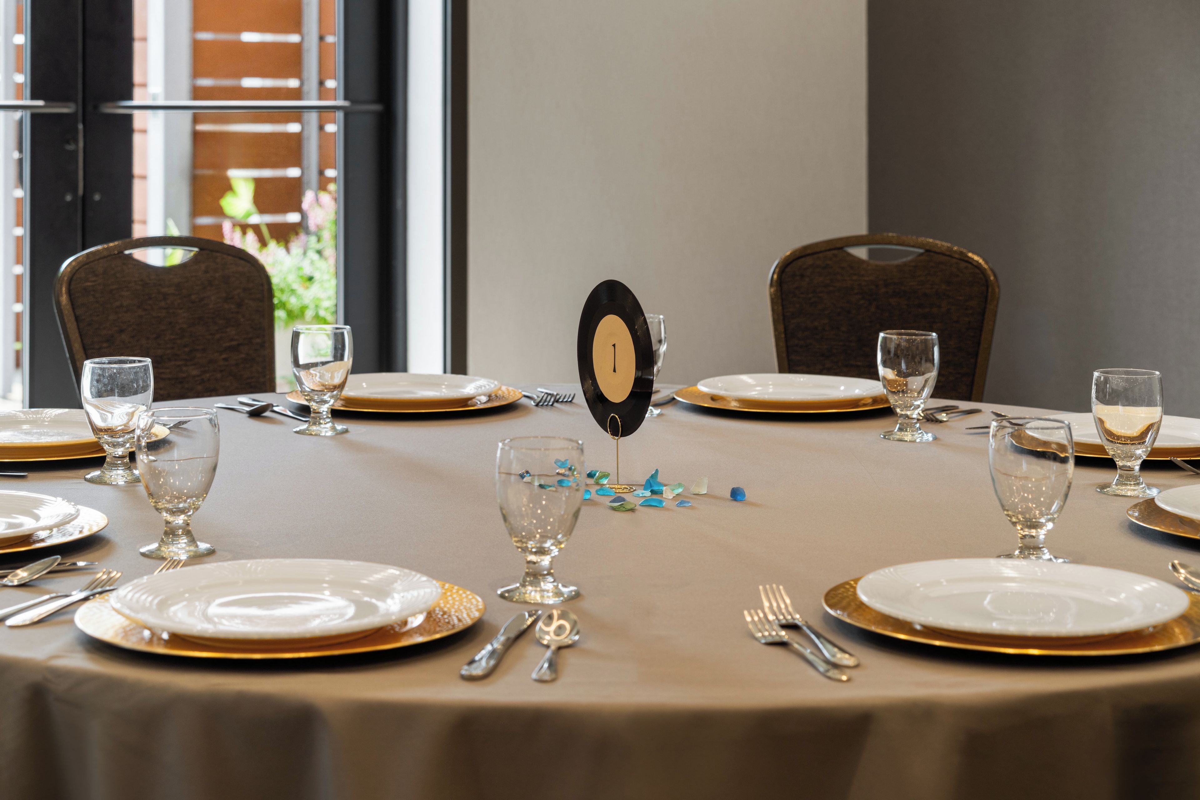 Una mesa redonda dispuesta para una comida, con platos numerados, copas transparentes y platos con detalles dorados.