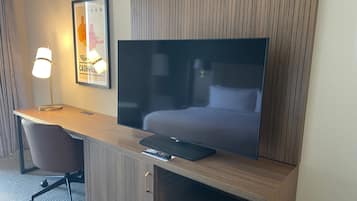 A hotel room with a large flat-screen TV on a wooden desk, a chair, a framed picture, and a lamp.