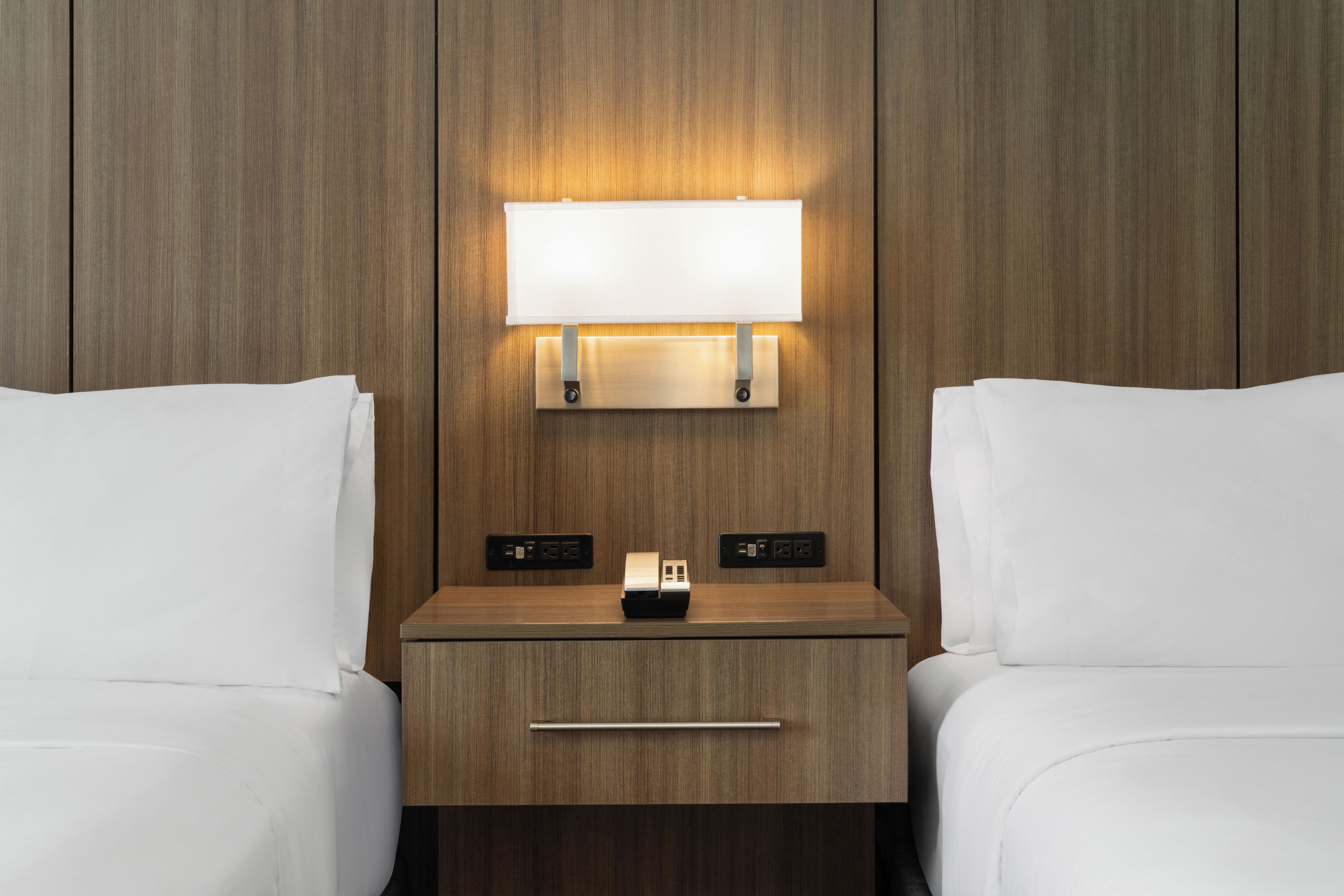 Ein Hotelzimmer mit einem hölzernen Kopfteil, einem Nachttisch mit Lampe und einer Steckdose.