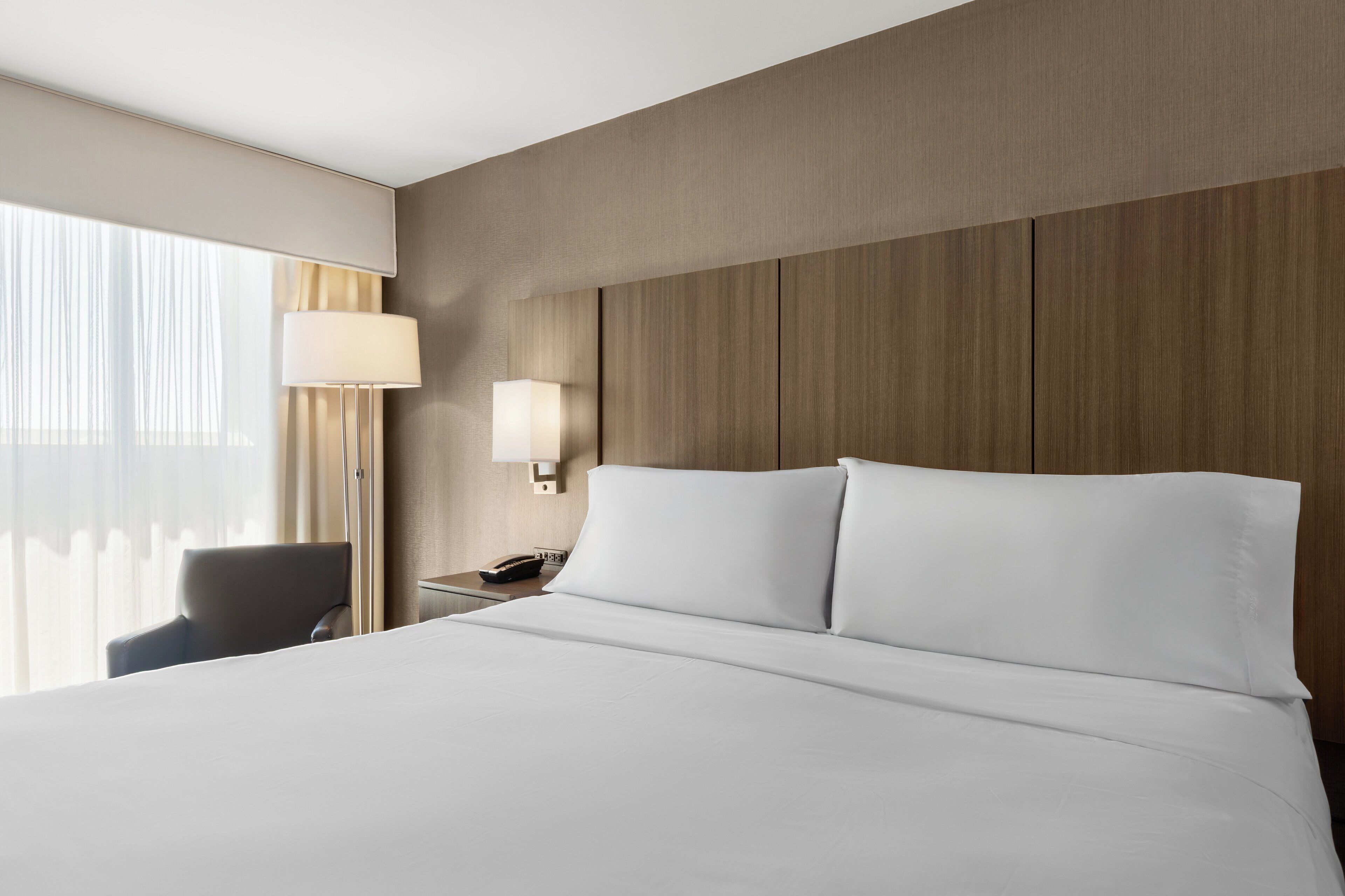 A hotel room with a large bed, a bedside table with a lamp, a chair, and a window with curtains.
