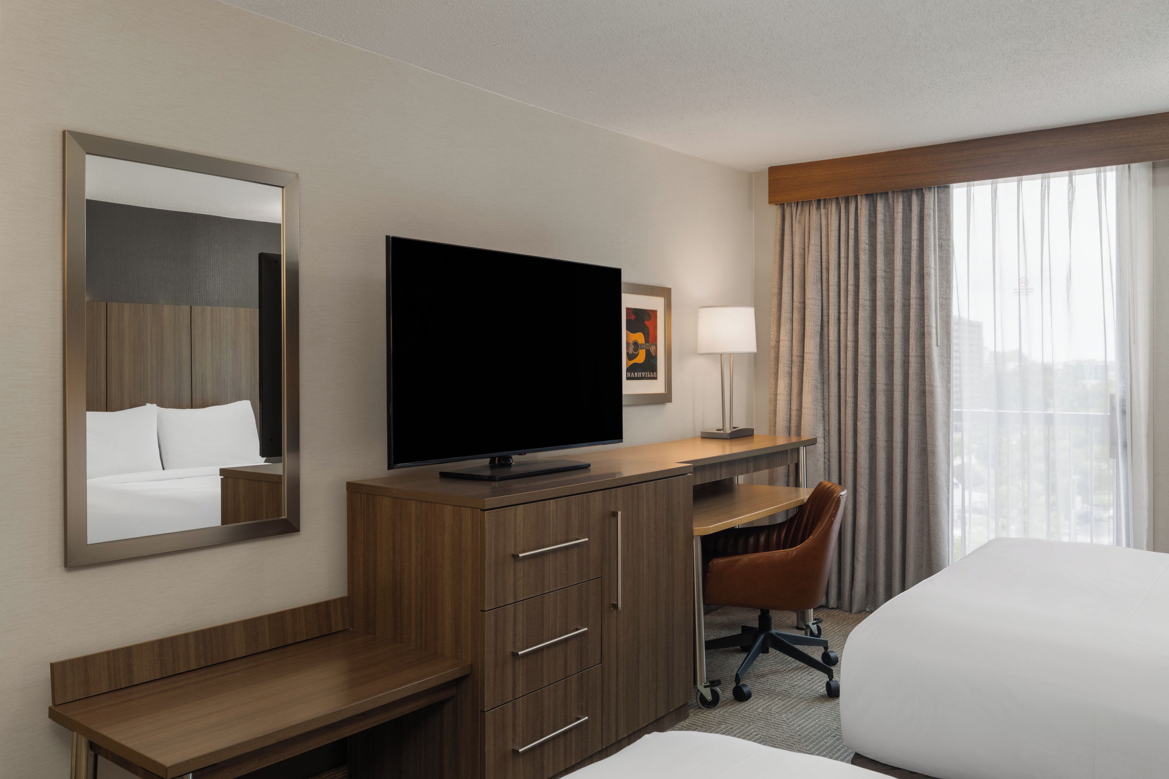 A hotel room with a bed, a desk with a flat-screen TV, a chair, and a large window with curtains.