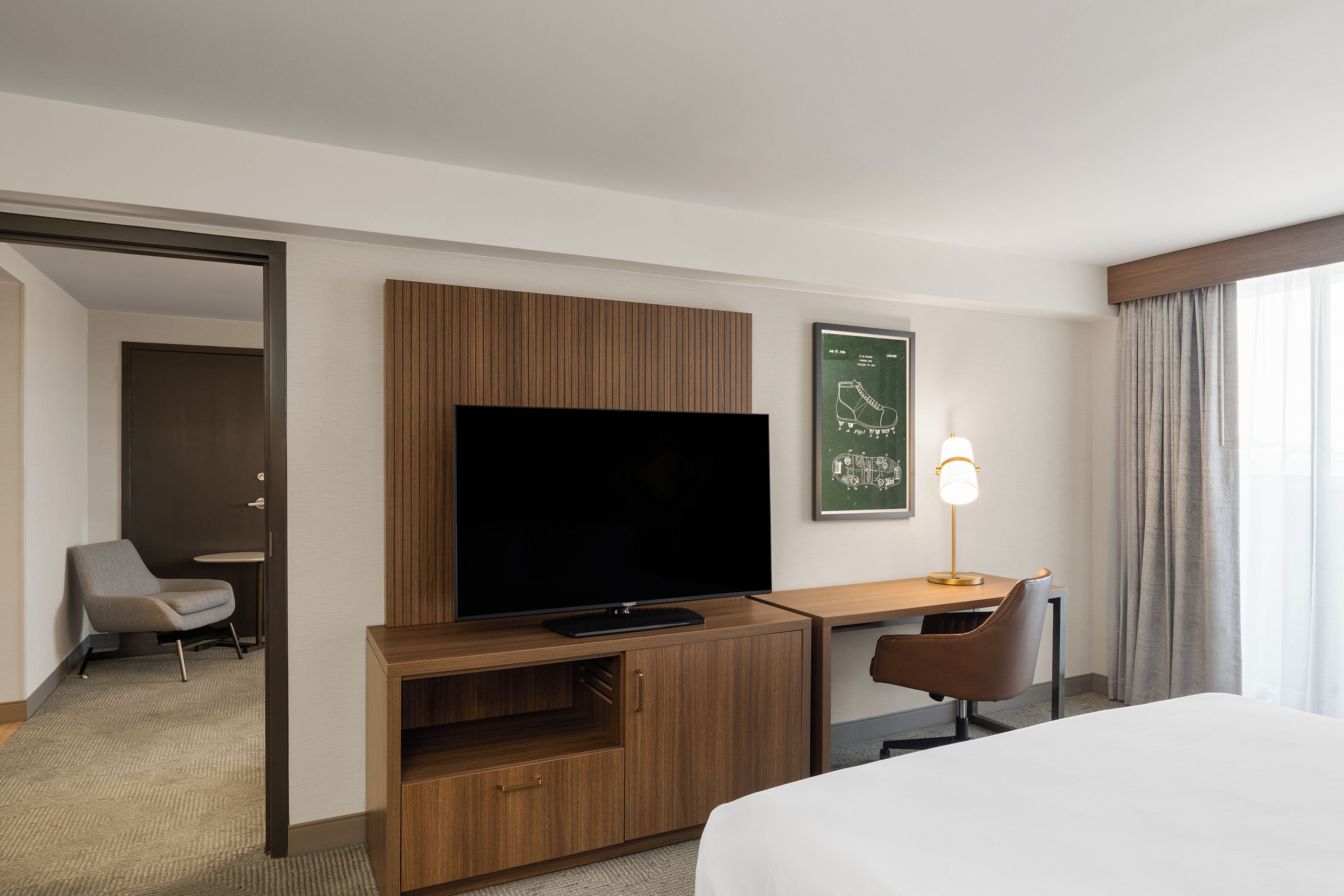 A hotel room with a large flat-screen TV, wooden furniture, a desk, a chair, and a bed.