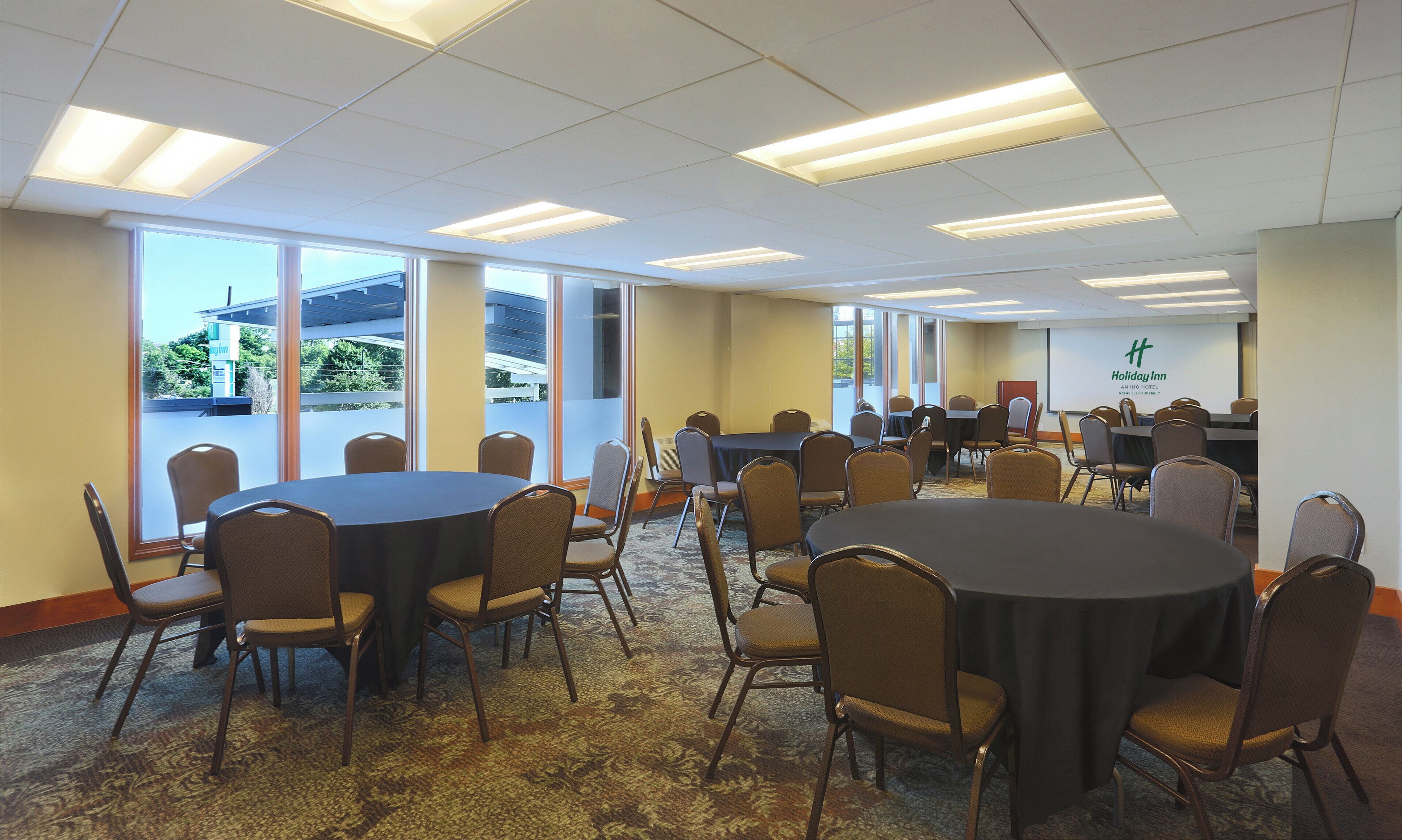 A conference room with multiple tables and chairs, large windows, and a Holiday Inn logo on the wall.