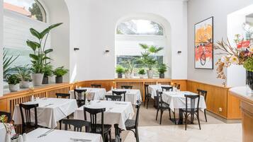 A restaurant with multiple tables and chairs, potted plants, and a view of the outside through large windows.