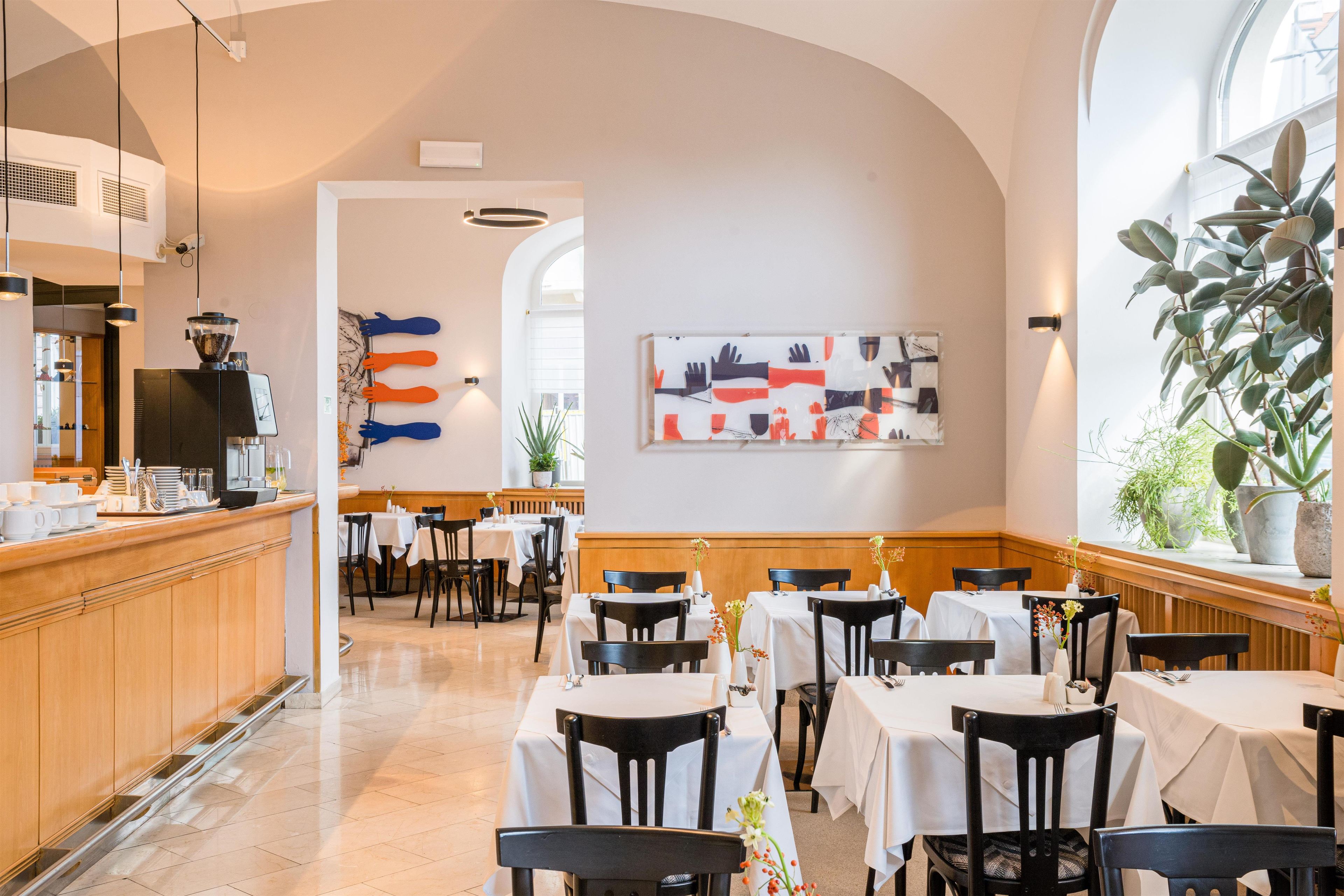 Un restaurante con una zona de bar, mesas con manteles blancos y un amplio ventanal con plantas.