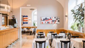 Un restaurante con una zona de bar, mesas con manteles blancos y un amplio ventanal con plantas.