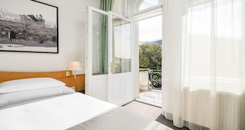 Una habitación de hotel con una cama, una mesita de noche, una lámpara, una puerta que da a un balcón y un cuadro en la pared.