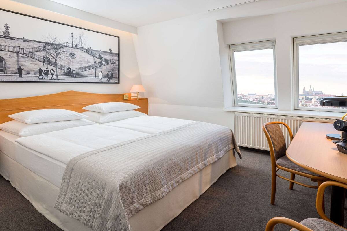 Hotelkamer met een groot bed, twee nachtkastjes, een bureau met stoel, een raam met uitzicht en een ingelijst kunstwerk aan de muur.