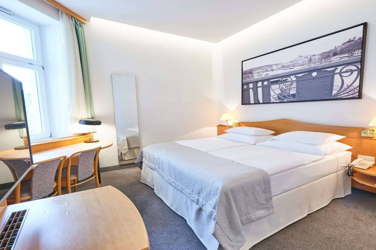 Een hotelkamer met een bed, nachtkastje, lamp, bureau, stoel en een groot raam.