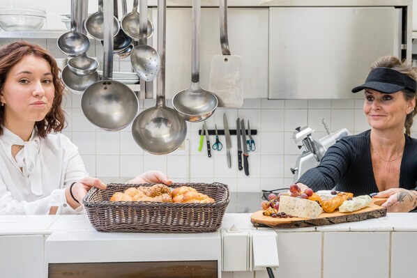 Una cocina con ollas y sartenes colgando, una cesta de pan y una tabla de quesos con uvas.