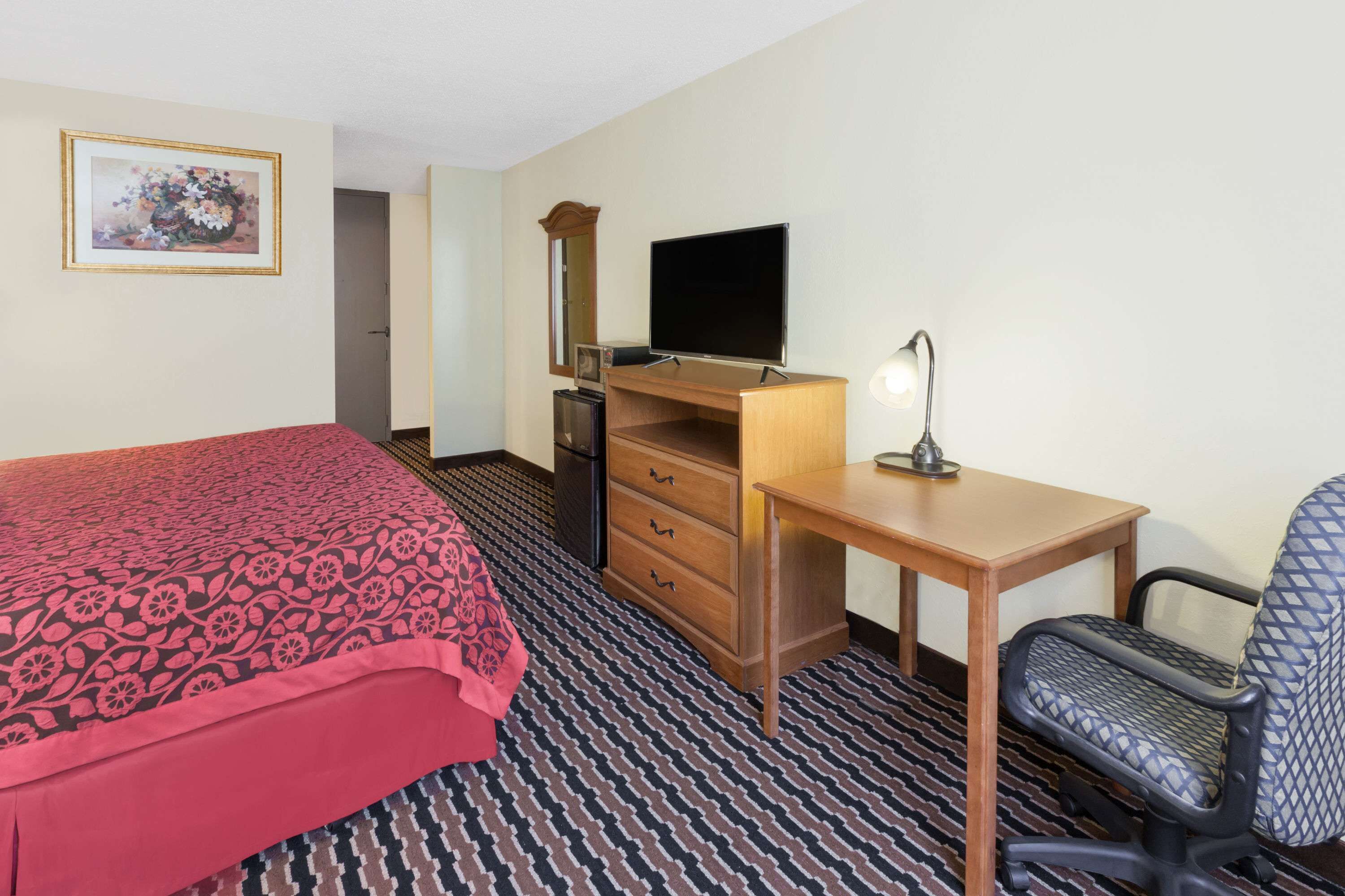 A hotel room with a bed, a desk with a chair, a television, a dresser, and a lamp.