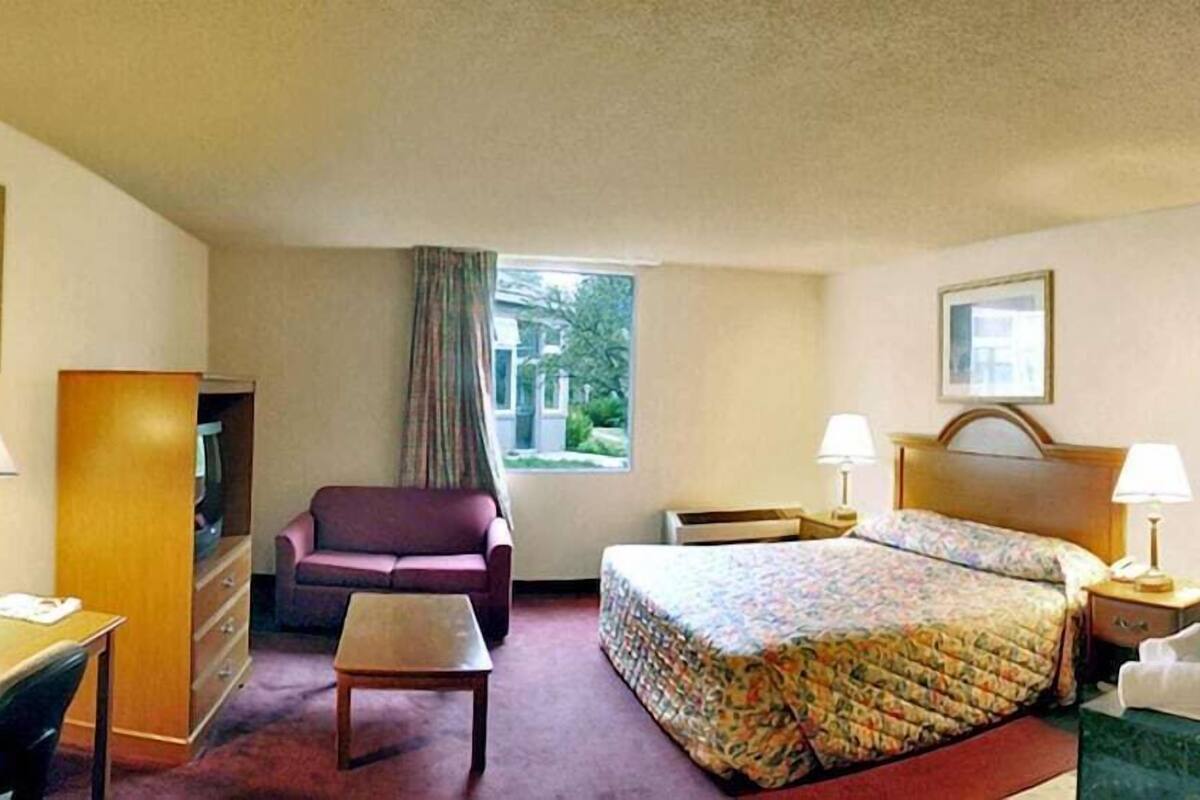A hotel room with a bed, a desk, a chair, a small table, a window with curtains, and a bathroom with a bathtub.