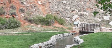 A stone-lined stream with a wooden bridge, surrounded by grass and rocky terrain.