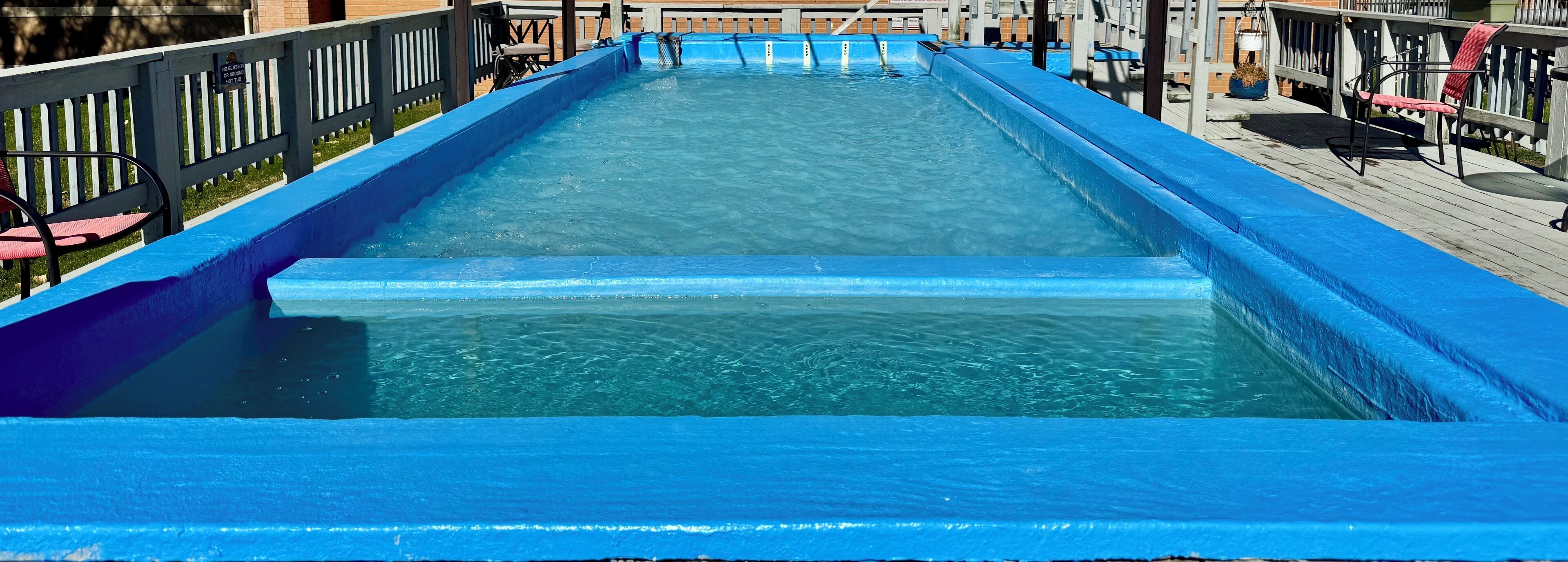 Ein langer, schmaler Swimmingpool mit blauer Umrandung, umgeben von einer Holzterrasse und einem weißen Zaun.