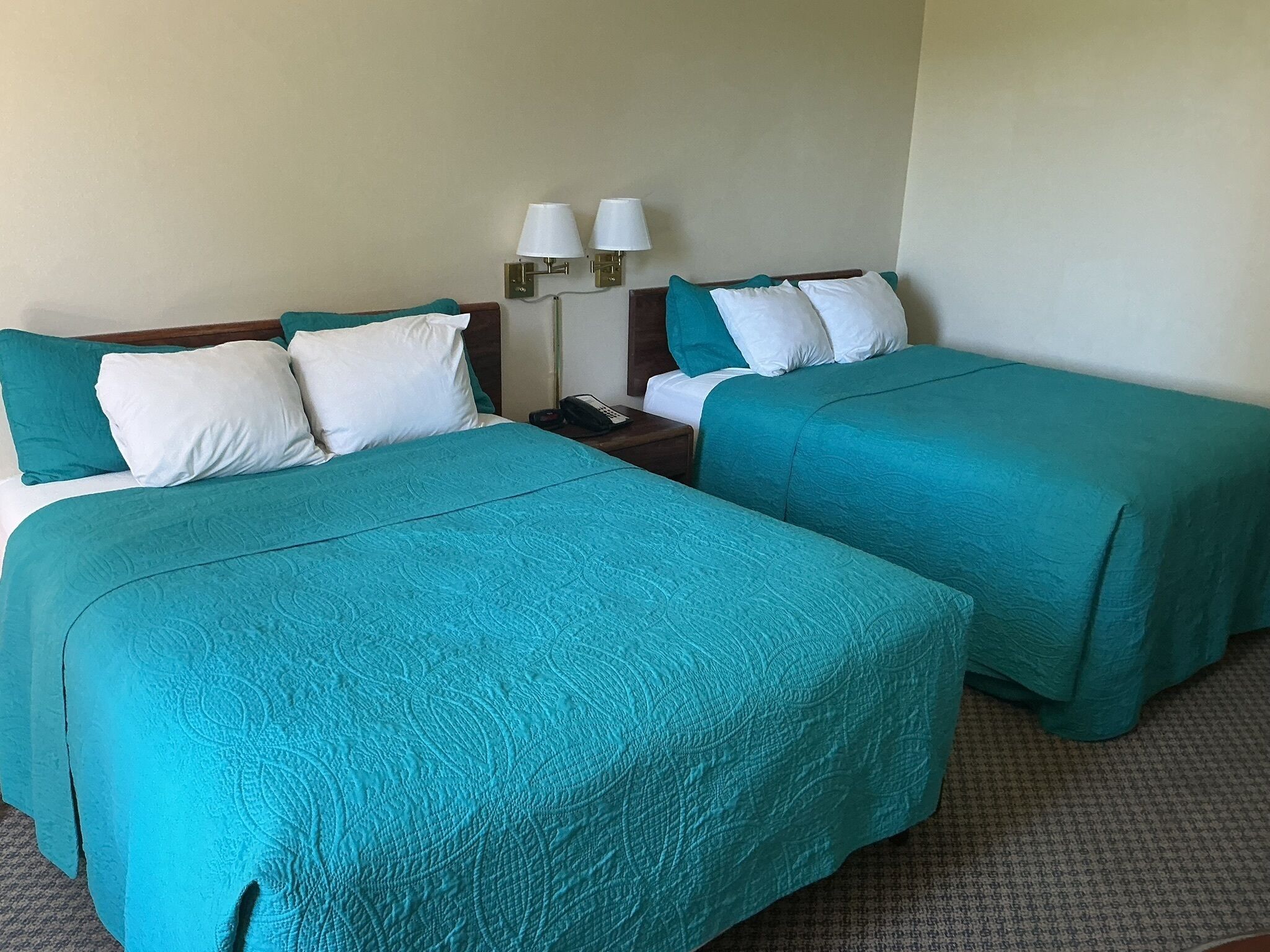 Habitación de hotel con dos camas, cada una con ropa de cama color aguamarina y almohadas blancas. Hay dos lámparas de noche y un teléfono en la mesita.
