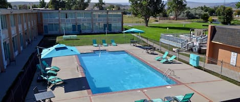 A swimming pool with lounge chairs and umbrellas, surrounded by a fenced area with a building in the background.