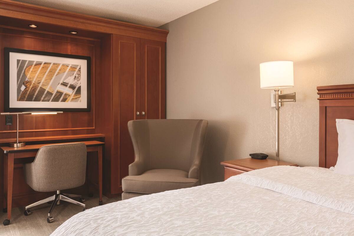 A hotel room with a bed, a desk, a chair, a framed picture on the wall, and a bedside lamp.