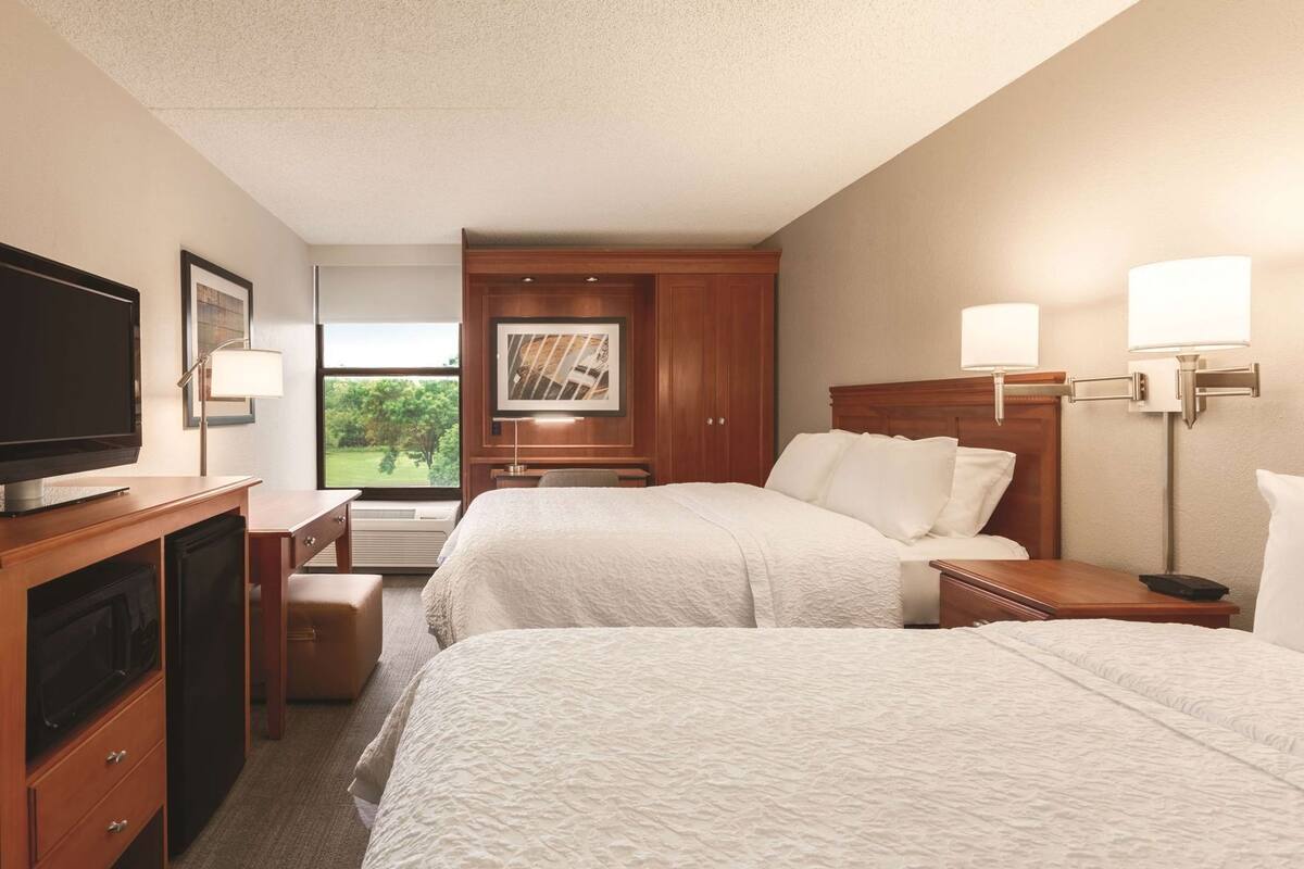 A hotel room with two beds, a TV, a desk, and a window with a view of greenery.