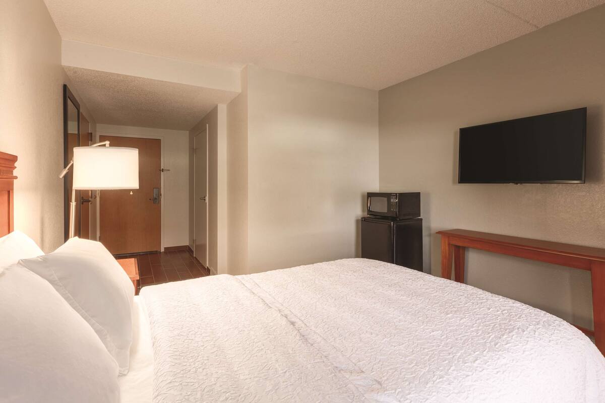 A hotel room with a large bed, a flat-screen TV mounted on the wall, a small refrigerator, and a wooden nightstand.