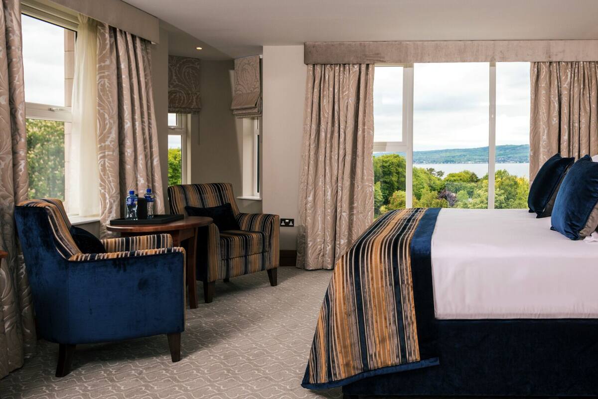 A hotel room with a large bed, two armchairs, a small table, and a view of a lake.
