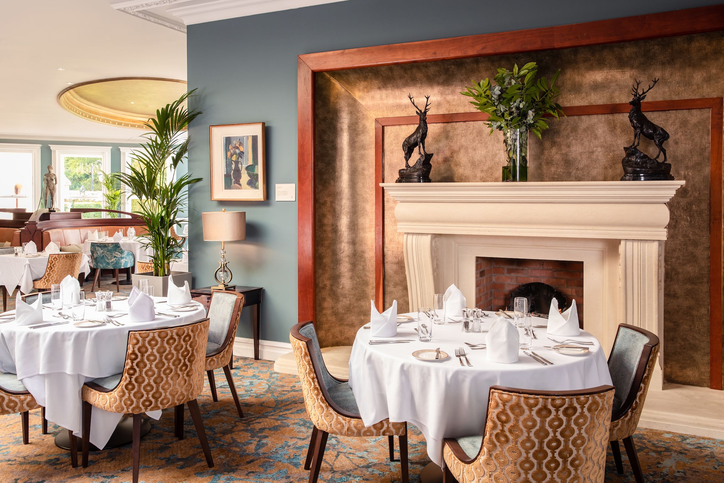 A dining area with round tables set for a meal, chairs with patterned upholstery, a fireplace with decorative deer sculptures, and a framed painting on the wall.