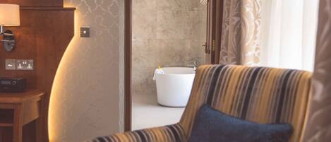 A hotel room with a striped armchair, a wooden side table with a lamp, a white bathtub, and a patterned wall.