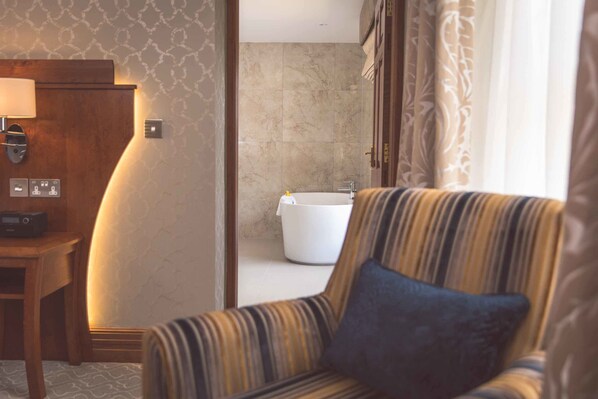 A hotel room with a striped armchair, a wooden side table with a lamp, a white bathtub, and a patterned wall.