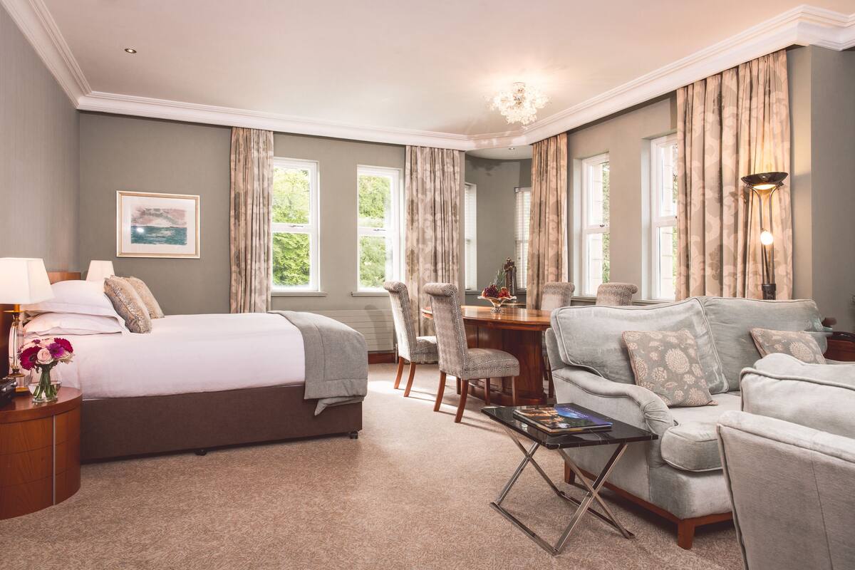A hotel room with a bed, a sofa, a dining table, and chairs. There are curtains on the windows, and a chandelier is visible.