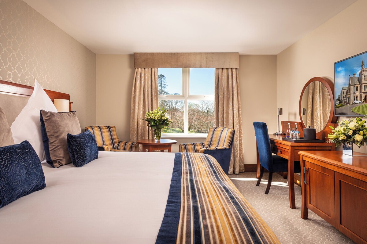 Une chambre d’hôtel avec un grand lit, un bureau avec une chaise, une petite table avec un vase et une fenêtre avec des rideaux.