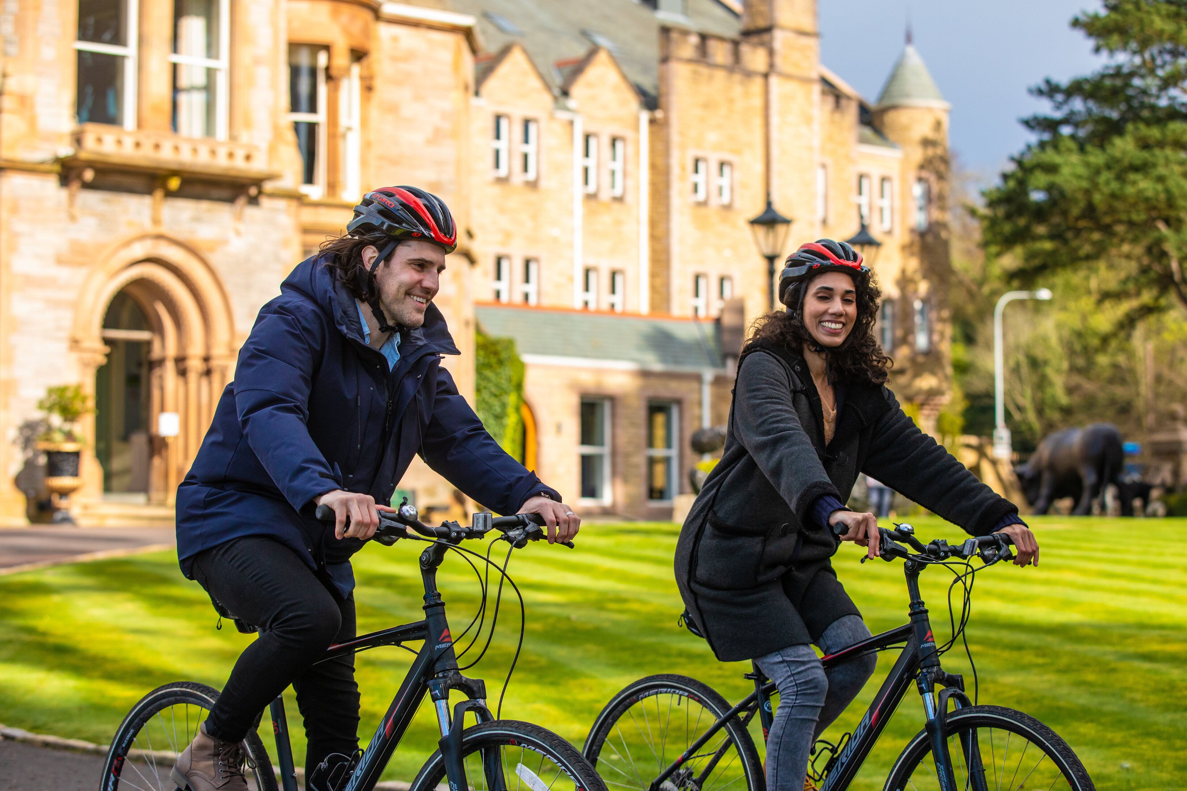 Two people riding bicycles in front of a historic building with a well-maintained lawn.