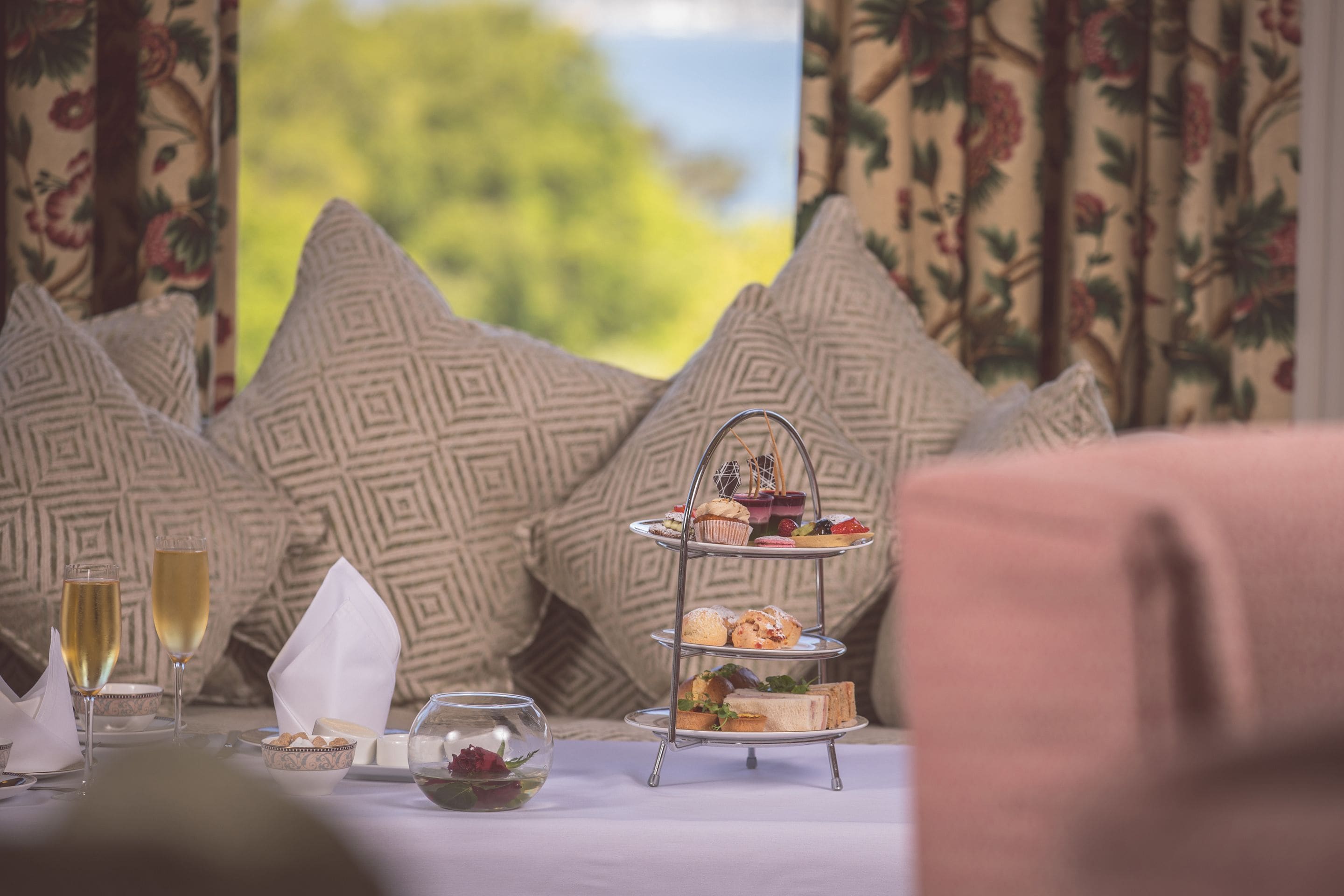 Un présentoir à trois niveaux garni de pâtisseries et de sandwichs, une coupe de champagne et un saladier de fruits sur une table, avec des coussins à motifs en arrière-plan.