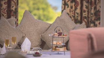 Un présentoir à trois niveaux garni de pâtisseries et de sandwichs, une coupe de champagne et un saladier de fruits sur une table, avec des coussins à motifs en arrière-plan.