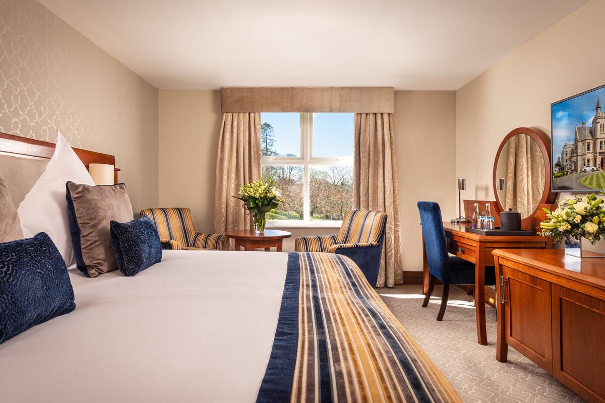A hotel room with a large bed, a desk, a chair, a small table with a vase of flowers, and a window with curtains.