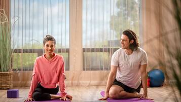 Dos personas sentadas sobre esterillas de yoga en una habitación con ventanas amplias y suelo de madera.