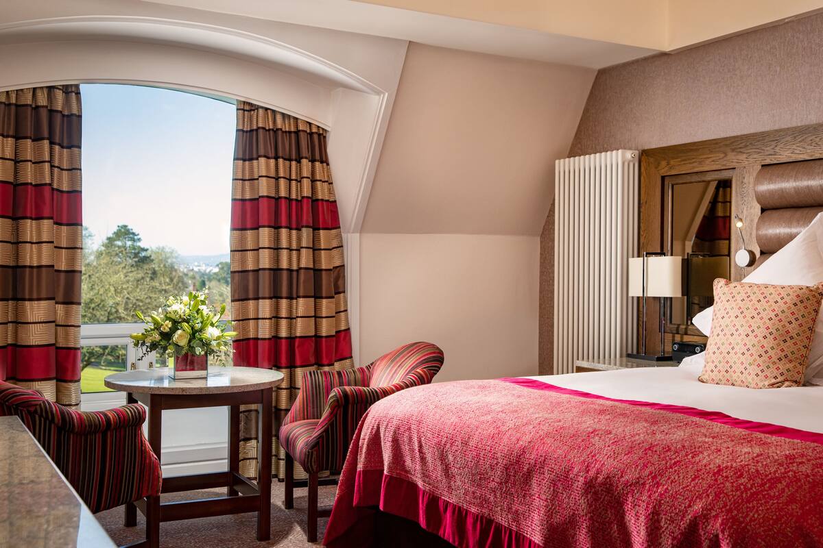 A hotel room with a bed, a round table with a vase of flowers, a striped armchair, and a view of the outdoors.