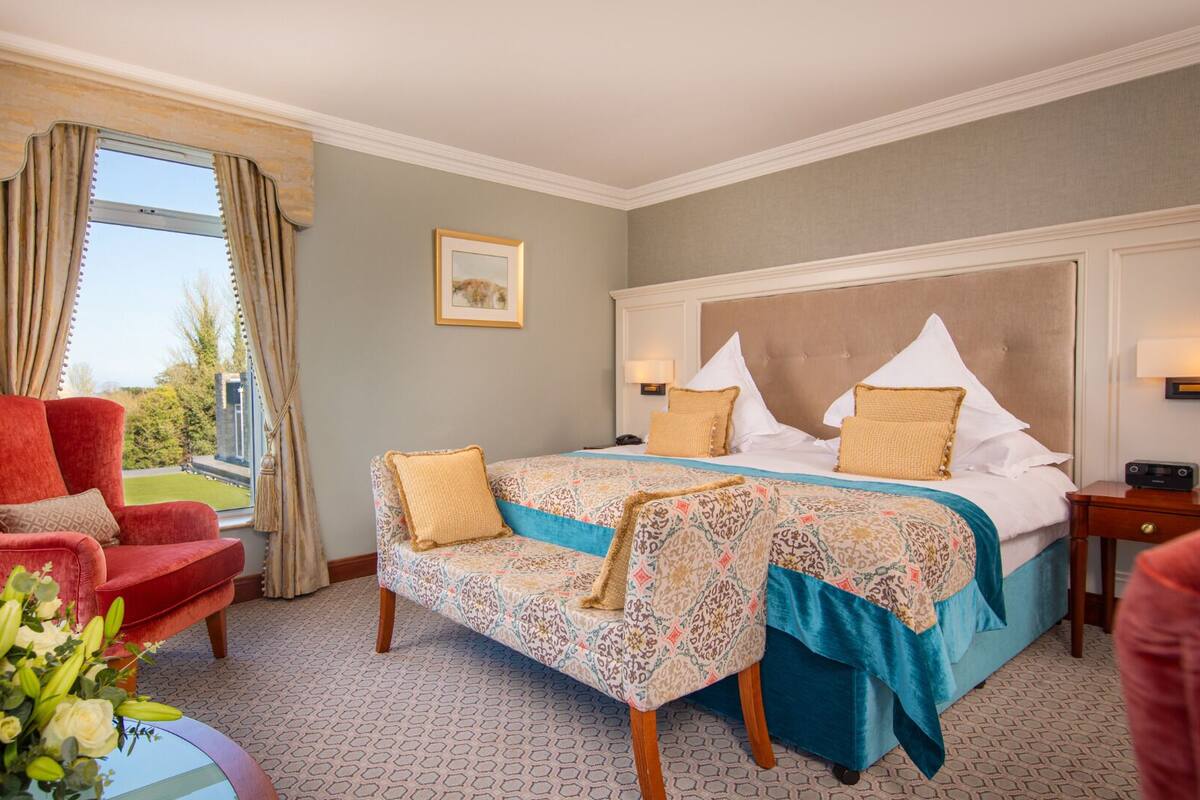 A hotel room with a large bed, a red armchair, a view of the outdoors, and a small table with a vase of flowers.