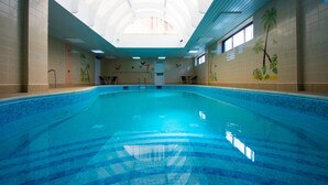 An indoor swimming pool with a tiled floor and walls, a high ceiling with skylights, and murals of palm trees and birds on the walls.